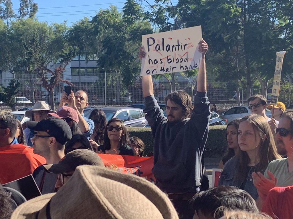 At protest against surveillance company Palantir in Palo Alto, a protestor holds up a sign that says "Palantir has blood on their hands"